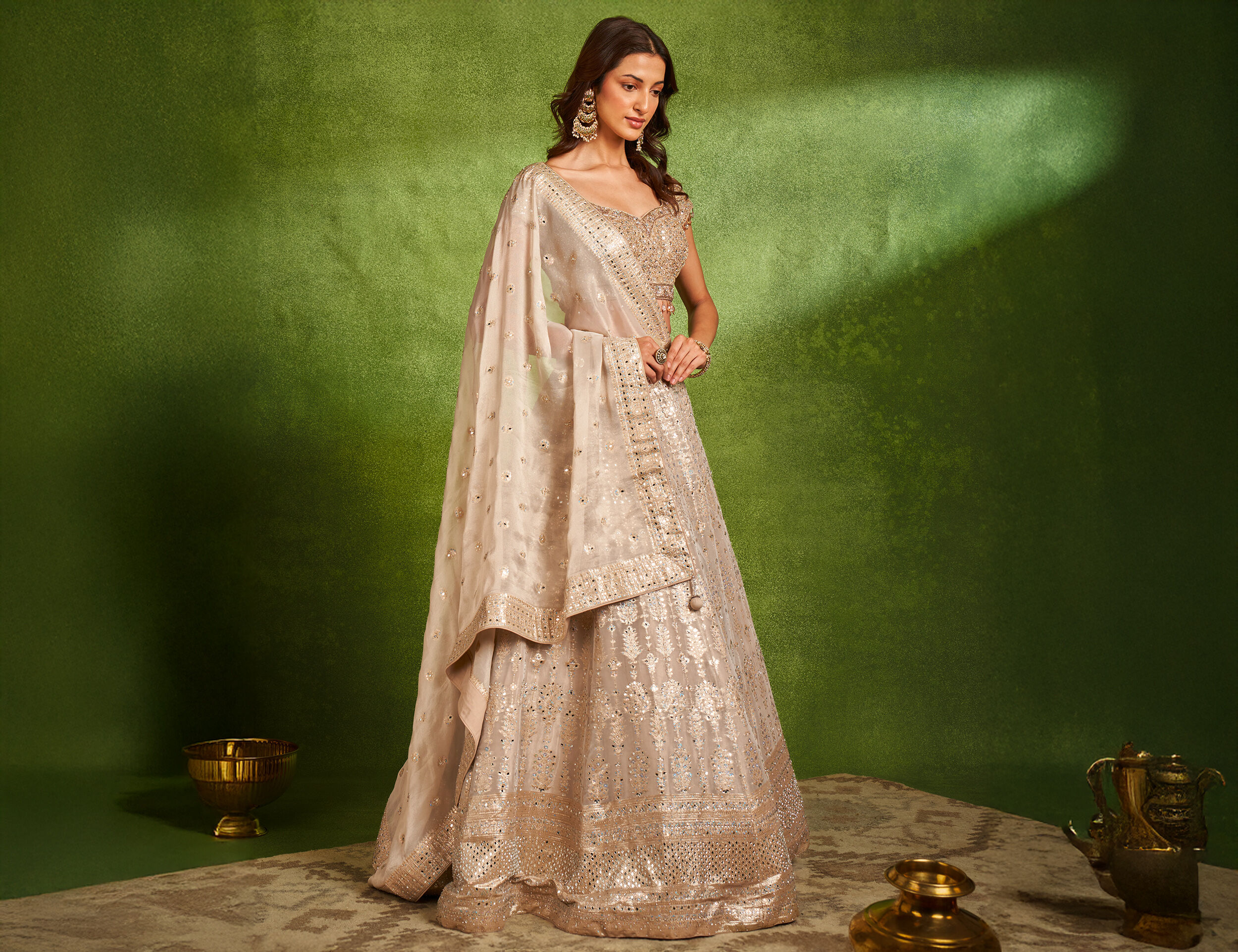 Elegant woman in a champagne gold embroidered lehenga with matching dupatta, styled with statement earrings, posing against a green festive backdrop for Navratri or wedding celebrations.